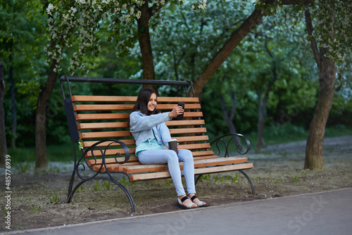 A young girl takes a selfie on her phone while sitting on a bench in a city park. Spring. Apple trees in bloom.  © Viktoriya
