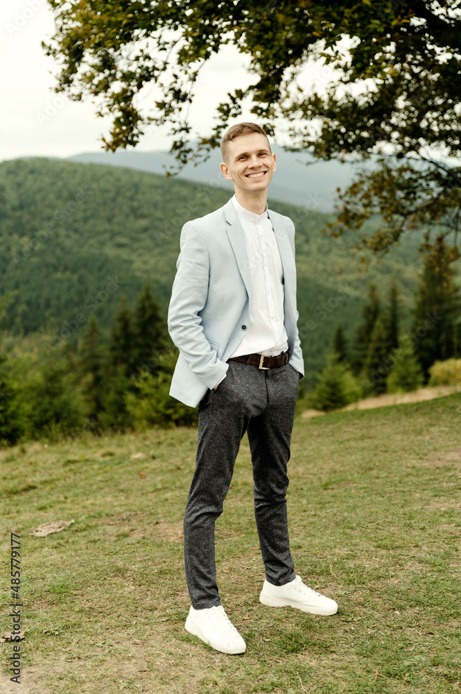 a young groom with a blue jacket stands near a tree against the backdrop of mountains a young groom with a blue jacket stands near a tree against the backdrop of mountains
