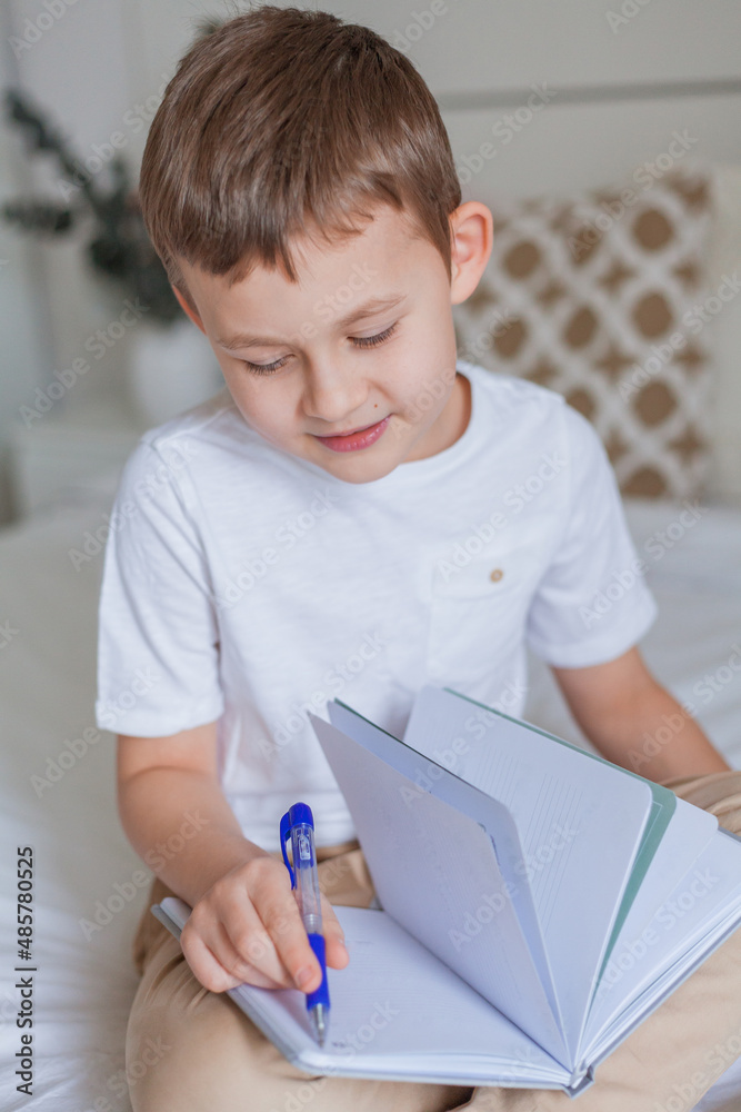 Cute schoolboy doing homework on the bed. A little boy of 7 years old is doing tasks online. Home schooling. Cute schoolboy doing homework on the bed. A little boy of 7 years old is doing tasks online. Home schooling.