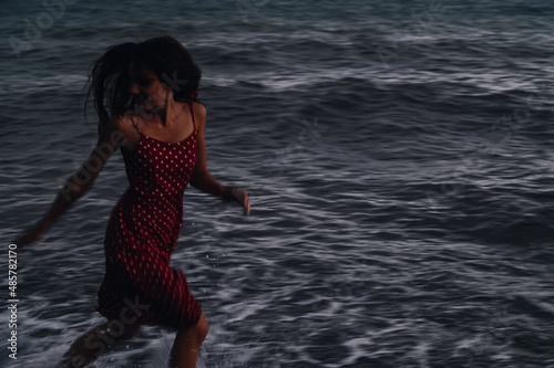 Fototapeta Naklejka Na Ścianę i Meble -  A young girl in a dress runs along the coastline on the waves on a summer evening.