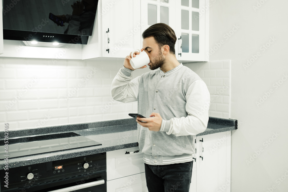 Young man is drinking coffee, the man with the phone in his hand is leaning on the counter and drinking coffee, modern kitchen and young man. Selective focus, noise effect.
