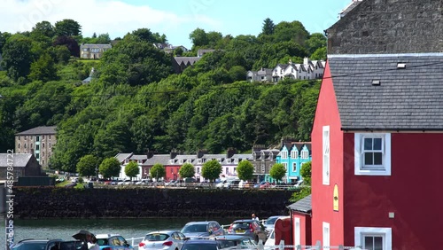 Several tourists walk along the promenade between parked cars in the coloured village of Tobermory on the Isle of Mull in Scotland. Medium shot photo