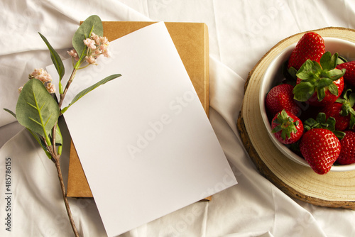 Blank notebook, flowers and strawberries on white sheet. Flat lay, top view