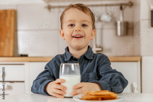 Little boy sitting in the kitchen and drinking milk. Fresh milk in glass, dairy healthy drink. Healthcare, source of calcium, lactose. Cozy and modern interior. Preschool child with casual clothing.