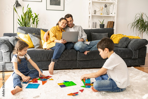 Happy family of four spends weekend at home. Parents using laptop sitting on the sofa in cozy living room, watching movies while two kids playing with constructor on the floor. Family idilia photo