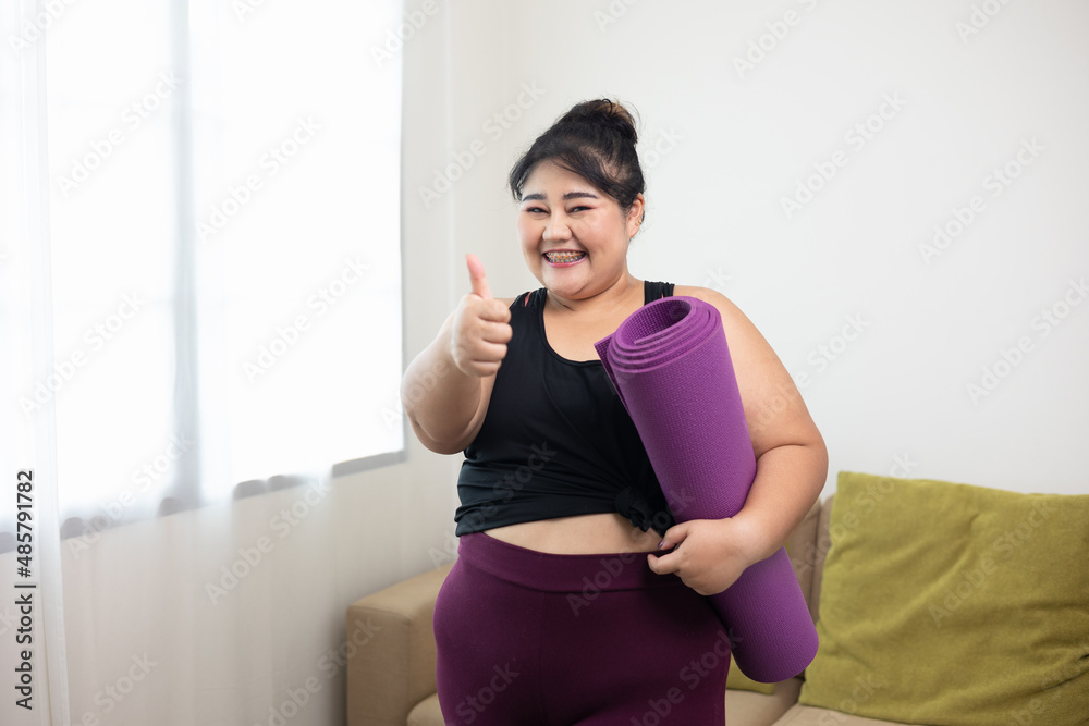 Attractive asian young fitness woman plus size holding yoga mat workout at home in living room. Female training and exercise wearing sport wear fit body. Happy and is proud of herself. Attractive asian young fitness woman plus size holding yoga mat workout at home in living room. Female training and exercise wearing sport wear fit body. Happy and is proud of herself.