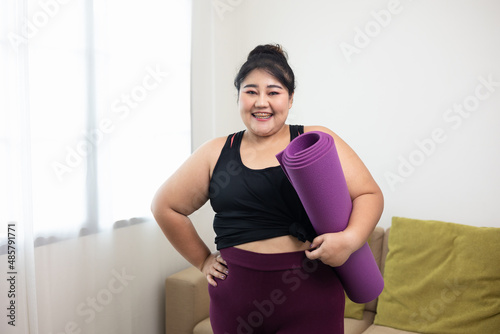 Attractive asian young fitness woman plus size holding yoga mat workout at home in living room. Female training and exercise wearing sport wear fit body. Happy and is proud of herself.
