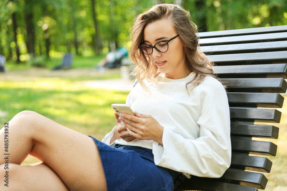 young woman holding phone and sitting on the bench in the summer park. girl reading sms message on her phone outdoors close up. young woman holding phone and sitting on the bench in the summer park. girl reading sms message on her phone outdoors close up.