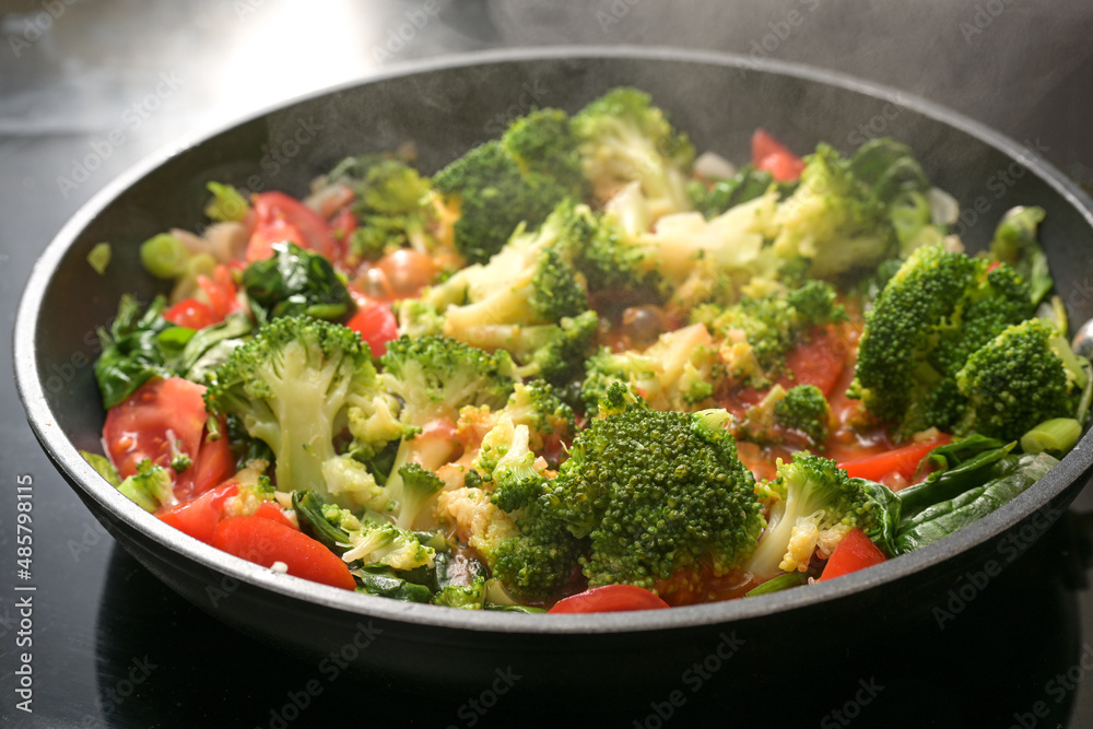 Steaming red and green vegetables in a pan with broccoli, tomatoes, spring onions and spinach on the stove, cooking vegetarian food for a healthy diet, selected focus