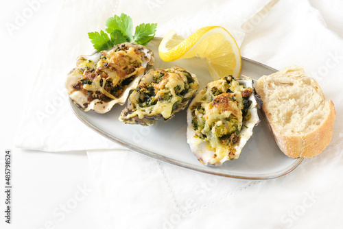 Three baked oysters Rockefeller with spinach and cheese on a small plate with lemon, bread and parsley garnish, festive appetizer on a white table, copy space, selected focus