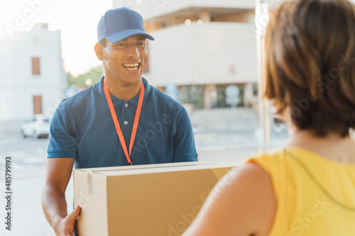 Billede på lærred Young, smiling Latino delivery man delivering a package at dusk to an unrecognizable woman at her front door