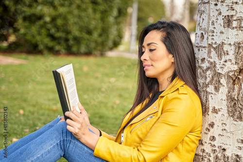Dark-haired woman sitting in the grass, leaning against a tree, reading a book on a spring day in a park. . High quality photo