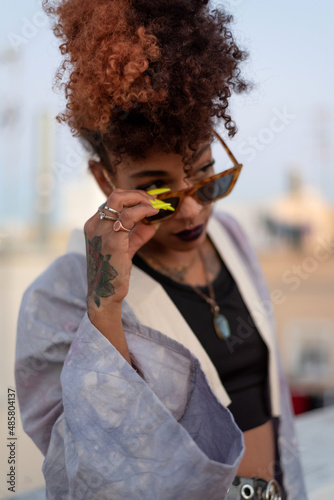 Stylish black female with curly hair and sunglasses wearing a lilac robe while resting on terrace against cloudless blue sky in Cadiz, Spain. photo