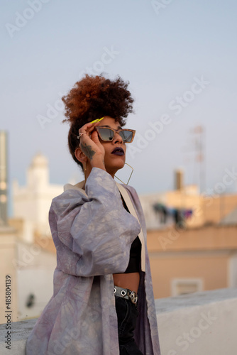 Stylish black female with curly hair and sunglasses wearing a lilac robe while resting on terrace against cloudless blue sky in Cadiz, Spain photo