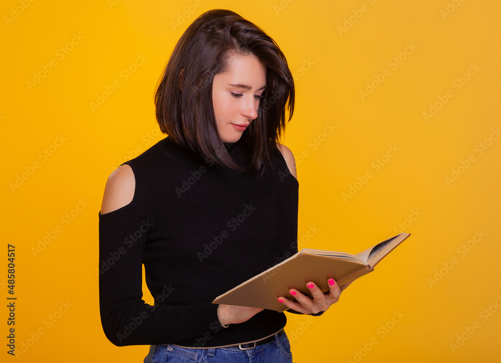 Pretty woman with a book in hands smiling at camera. Clever and excited lady holding open book. Studio shot isolated on yellow background