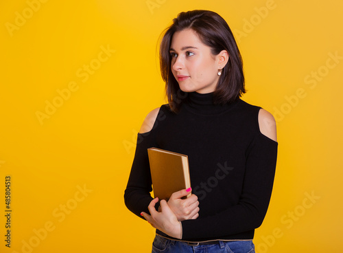 Pretty woman with a book in hands smiling at camera. Clever and excited lady embrace book. Studio shot isolated on yellow background