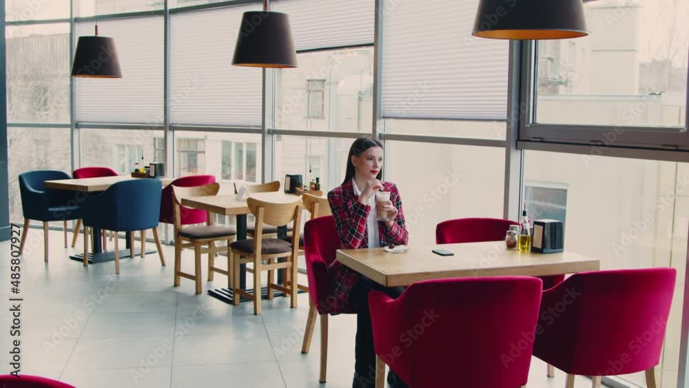 Young business woman is sitting at table in cafe and drinking coffee, enjoying her time off, tasting favorite beverage and looking at window, taking a pause in her busy day, Zoom in, Slow motion.