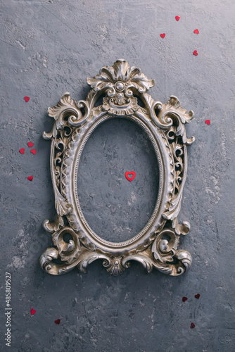 A concrete gray background with a vintage silver photo frame, in which a photo of young people in love is inserted, next to it lies a scattering of red sequins in the shape of a heart.