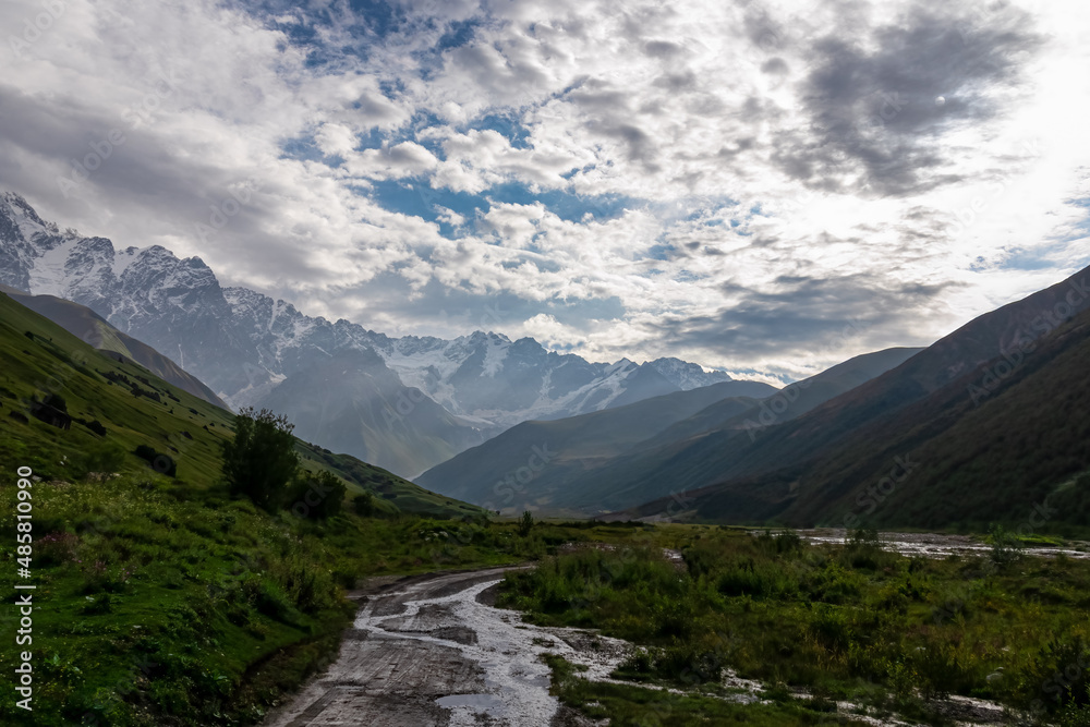 A hiking path leading to the Shkhara Glacier in the Greater Caucasus Mountain Range in Georgia, Svaneti Region, Ushguli. Snow-capped mountains in the back. Wanderlust. Ponds on the road from rain