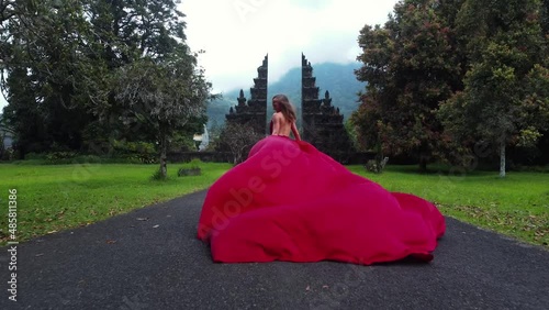 Pretty asian woman runs to balinese gates in long red dress and drone follows in slowmotion on Hangara gates in Bali Indonesia. fashion concept. High quality fullhd aerial footage