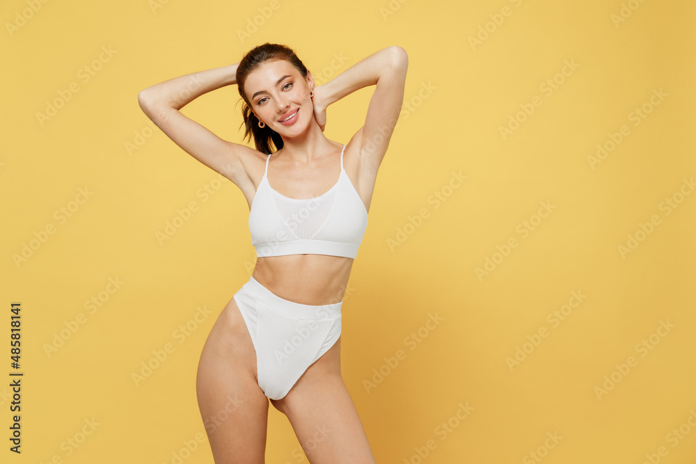 Smiling lovely attractive young brunette woman 20s wearing white underwear with perfect fit body standing ties hair in ponytail look camera isolated on plain yellow color background studio portrait - obrazy, fototapety, plakaty 