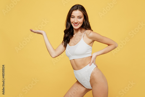 Smiling lovely satisfied young brunette woman 20s in white underwear with perfect fit body standing posing hold show empty arm with workspace area isolated on plain yellow background studio portrait