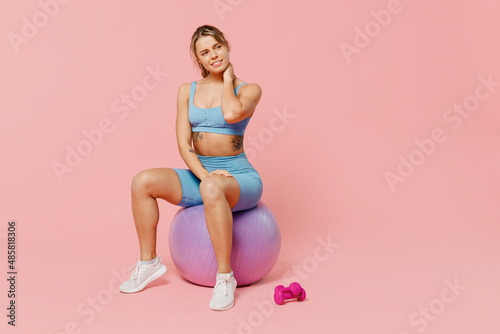 Full size young sporty fitness trainer instructor woman wear blue tracksuit spend time in home gym sit on fitball hold neck suffer from pain isolated on plain pink background. Workout sport concept.