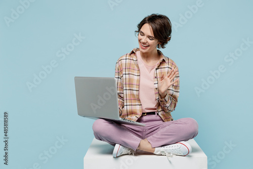 Full body young smiling cheerful happy woman 20s wear brown shirt sit on white chair hold use work on laptop pc computer waving hand speak by video call isolated on pastel plain light blue background. photo