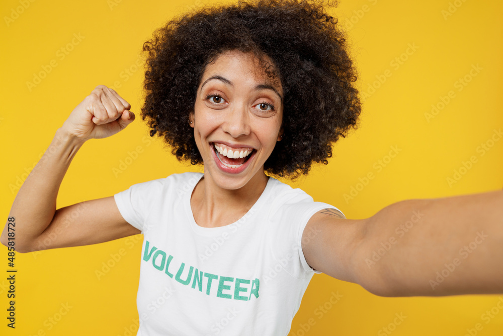 Close up young woman of African American ethnicity wears white volunteer t-shirt do selfie shot pov on mobile phone do winner gesture isolated on plain yellow background. Voluntary free help concept