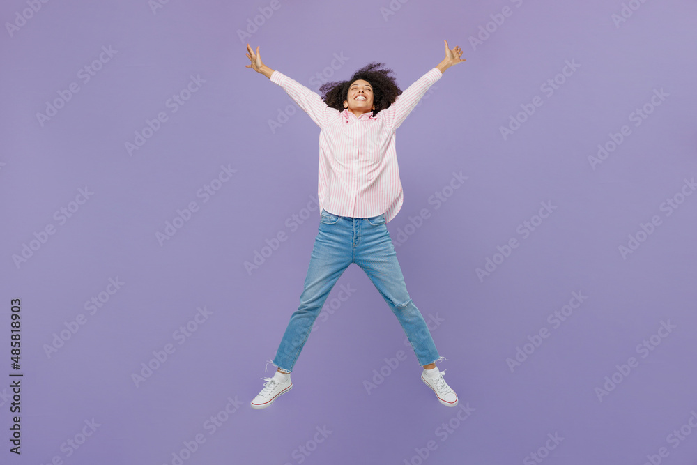Full body exultant jubilant happy excited fun young woman of African American ethnicity wear pink striped shirt jump high with outstretched hands legs isolated on plain pastel light purple background.