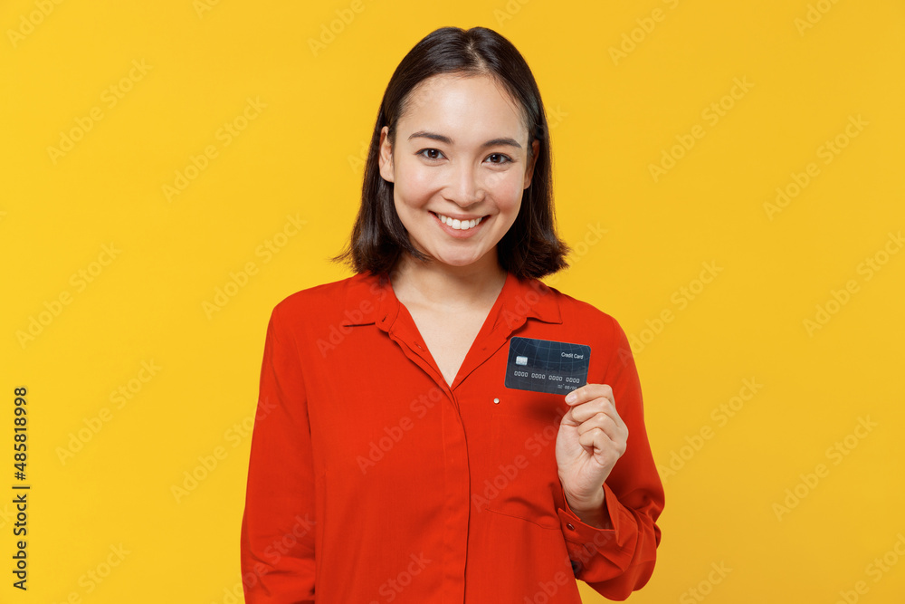 Satisfied smiling amazing vivid happy young woman of Asian ethnicity 20s years old wears orange shirt looking camera hold in hand credit bank card isolated on plain yellow background studio portrait. Satisfied smiling amazing vivid happy young woman of Asian ethnicity 20s years old wears orange shirt looking camera hold in hand credit bank card isolated on plain yellow background studio portrait.