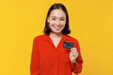 Satisfied smiling amazing vivid happy young woman of Asian ethnicity 20s years old wears orange shirt looking camera hold in hand credit bank card isolated on plain yellow background studio portrait.