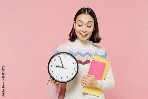 Smiling happy teen student girl of Asian ethnicity wear sweater backpack hold books look at clock isolated on pastel plain light pink background Education in high school university college concept