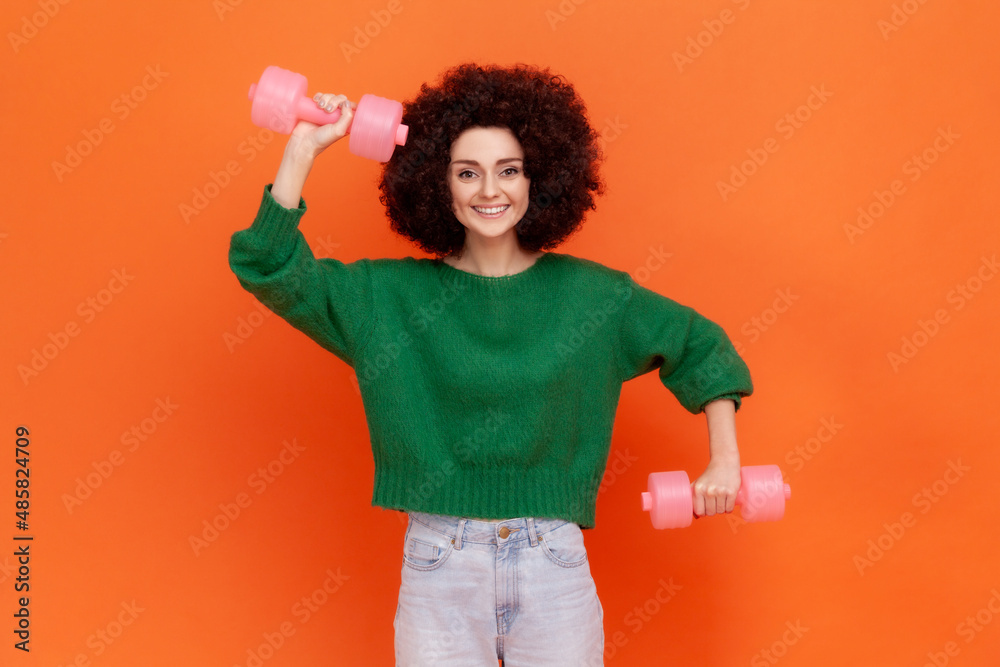 Happy positive sporty woman with Afro hairstyle wearing green casual style sweater doing exercises for biceps, using dumbbells, health care. Indoor studio shot isolated on orange background.