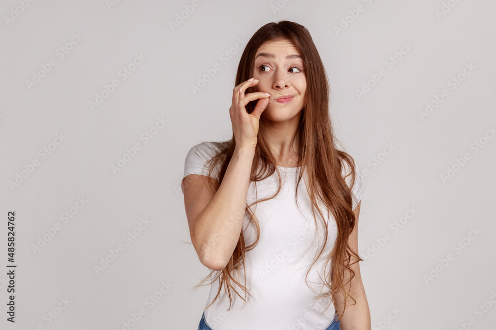 Attractive woman with long dark hair and mystery look making zip gesture to close mouth, keeping secret, zipping lips, wearing white T-shirt. Indoor studio shot isolated on gray background. Attractive woman with long dark hair and mystery look making zip gesture to close mouth, keeping secret, zipping lips, wearing white T-shirt. Indoor studio shot isolated on gray background.