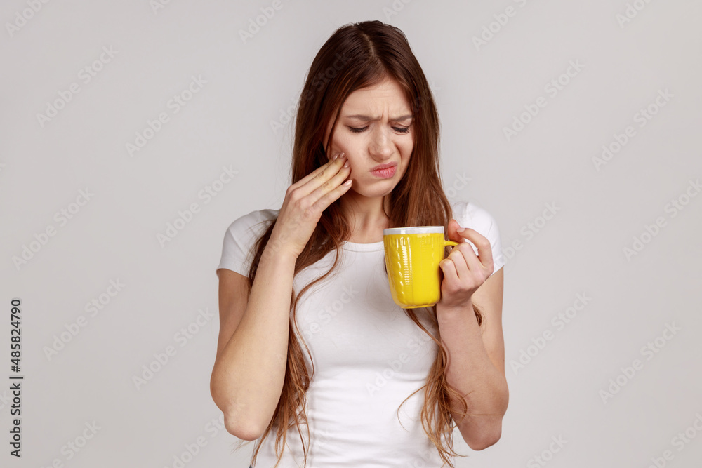 Sick unhealthy woman touching chin suffering tooth ache, has sensitive teeth, feels pain after drinking cold water, wearing white T-shirt. Indoor studio shot isolated on gray background. Sick unhealthy woman touching chin suffering tooth ache, has sensitive teeth, feels pain after drinking cold water, wearing white T-shirt. Indoor studio shot isolated on gray background.
