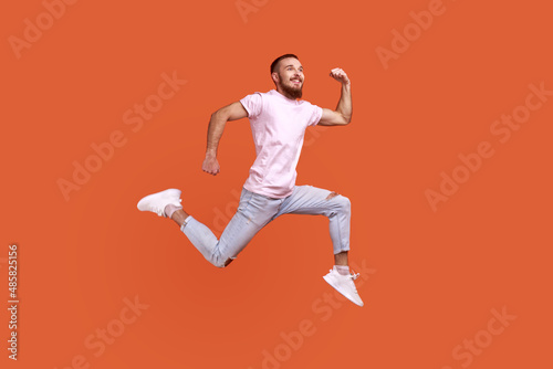 Full length portrait of positive inspired bearded man jumping in air or running quickly fast, expressing happiness, wearing pink T-shirt. Indoor studio shot isolated on orange background.