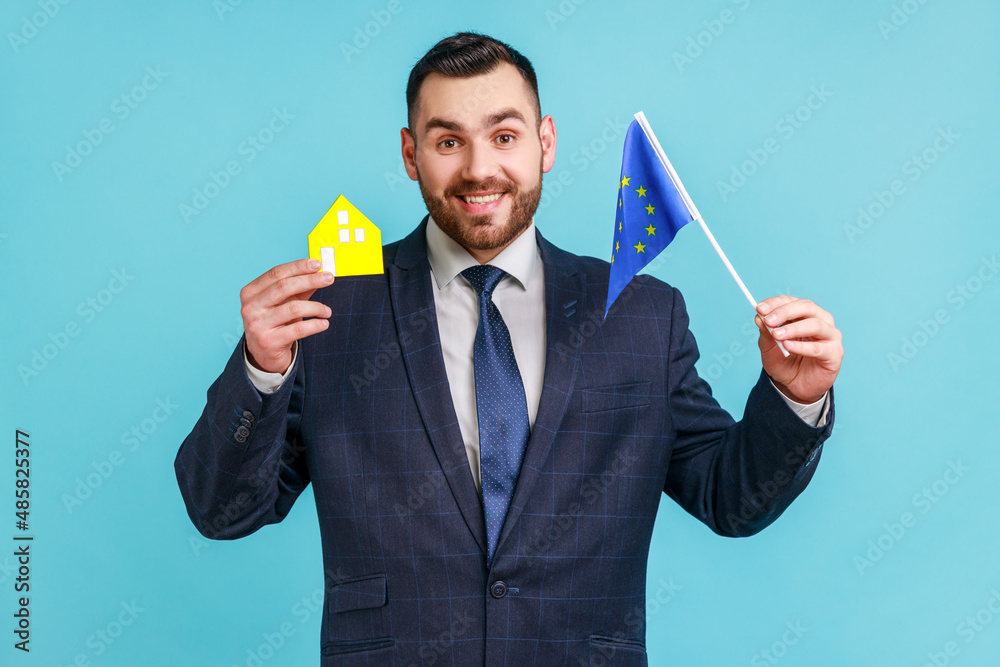 Good looking man with beard wearing official style suit holding model of paper house and europe union flag, dreaming about living in European country. Indoor studio shot isolated on blue background. Good looking man with beard wearing official style suit holding model of paper house and europe union flag, dreaming about living in European country. Indoor studio shot isolated on blue background.