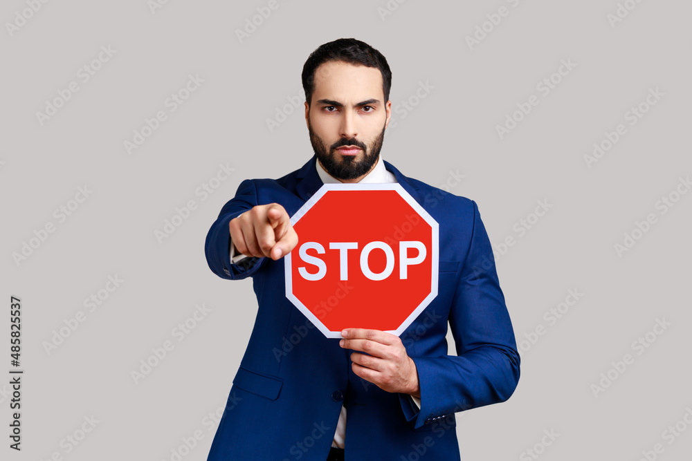 Serious bearded man pointing to camera and holding Stop road traffic sign as symbol of prohibition, restrictions, wearing official style suit. Indoor studio shot isolated on gray background.