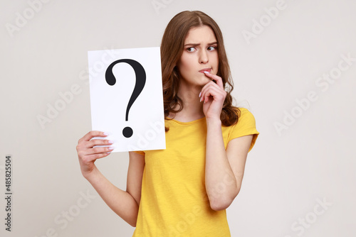 Young pensive woman in casual style attire holding paper with question mark over, thoughtful, face thinking about question, very confused idea. Indoor studio shot isolated on gray background.