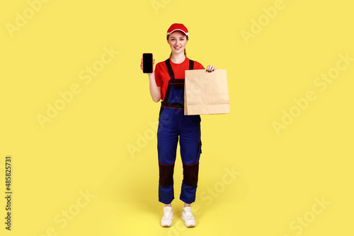 Full length portrait of friendly courier woman holding paper parcel and smart phone with empty display for advertisement, wearing overalls and red cap. Indoor studio shot isolated on yellow background