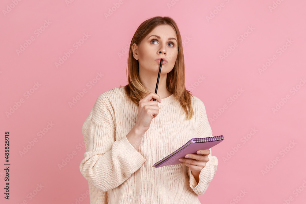 Portrait of pensive blond woman holding pen and paper notebook, being deep in thoughts, writing a composition, wearing white sweater. Indoor studio shot isolated on pink background. Portrait of pensive blond woman holding pen and paper notebook, being deep in thoughts, writing a composition, wearing white sweater. Indoor studio shot isolated on pink background.