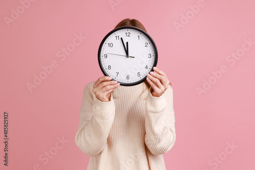 Portrait of blond woman holding wall clock hiding her face, time management, schedule and meeting appointment, wearing white sweater. Indoor studio shot isolated on pink background. photo
