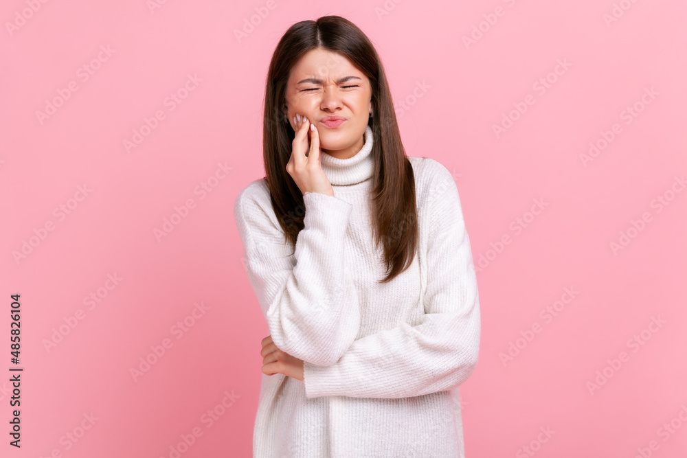 Unhealthy girl touching cheek, closing eyes with expression of terrible suffer from dental problems, wearing white casual style sweater. Indoor studio shot isolated on pink background. Unhealthy girl touching cheek, closing eyes with expression of terrible suffer from dental problems, wearing white casual style sweater. Indoor studio shot isolated on pink background.