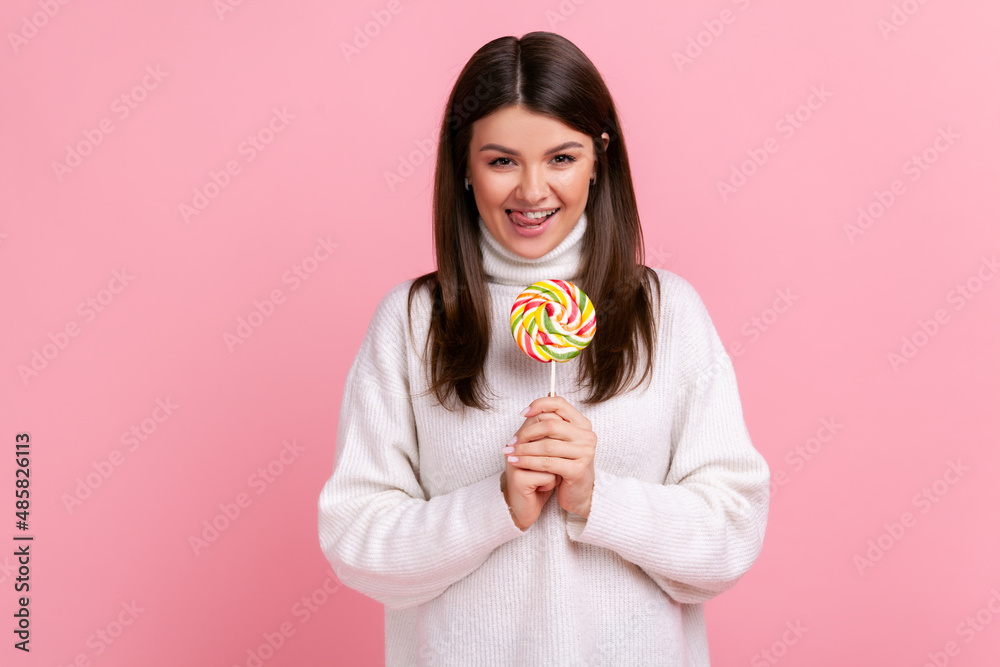 Portrait of hungry brunette female holding big tasty candy, looking at camera, showing tongue out, wearing white casual style sweater. Indoor studio shot isolated on pink background.