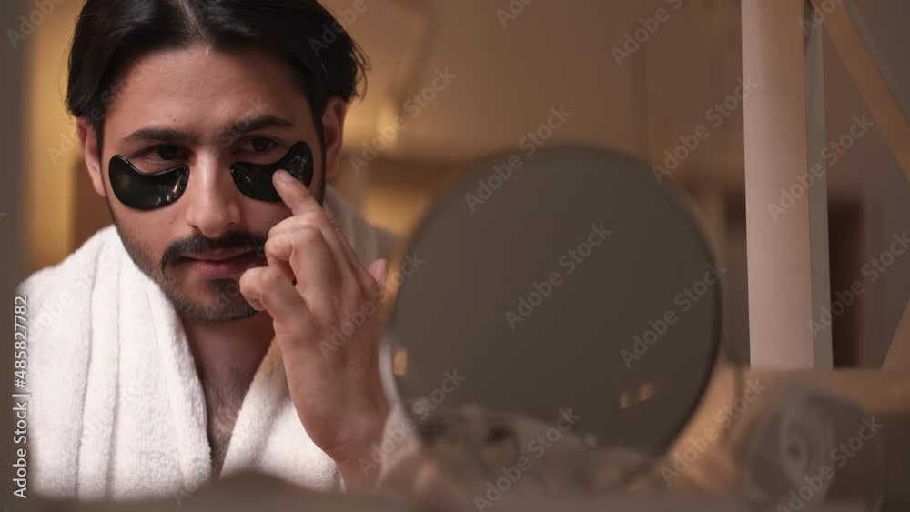 Male beauty care. Grooming routine. Rejuvenation procedure. Handsome man applying under eye silicone pads looking mirror light shadow bathroom interior.