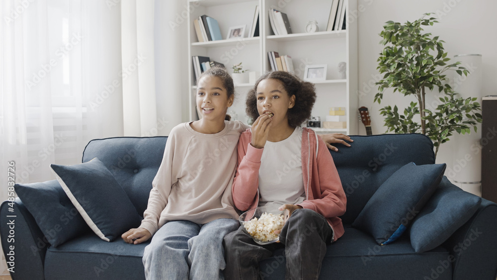 Two teen girls watching movie together, eating popcorn on the sofa, having fun Two teen girls watching movie together, eating popcorn on the sofa, having fun
