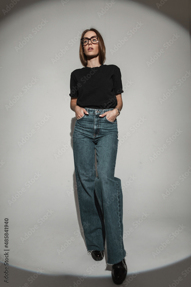 Stylish beautiful sexy woman model in fashionable clothes with a black  T-shirt and bell-bottomed jeans. Glamour pretty girl with glasses in  vintage outfit in the studio Photos | Adobe Stock