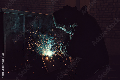 Side view of welder at work. Worker welds metal parts. Welding process, sparks, flame, smoke. 