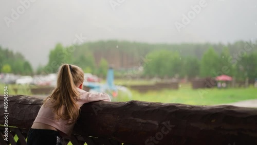 Bored little child leans on large old log railing and looks at helicopter with rotating propeller on terrace of rural cafe on rainy day slow motion photo